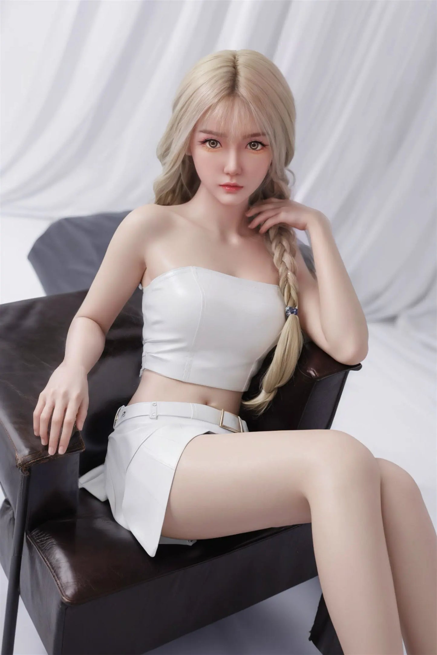 Adorey sex doll cool composed direct stare, warm amber-brown eyes, long blonde hair with single side braid, coral lips, small gold drop earring, white structured leather strapless crop top with seam detail straining over full round breasts with natural contour, sculpted toned bare midriff, white high-waist leather mini skirt with belt buckle detail, one hand raised delicately touching braid, other hand resting on armchair, seated on dark brown leather armchair with soft white draped curtain background – thr