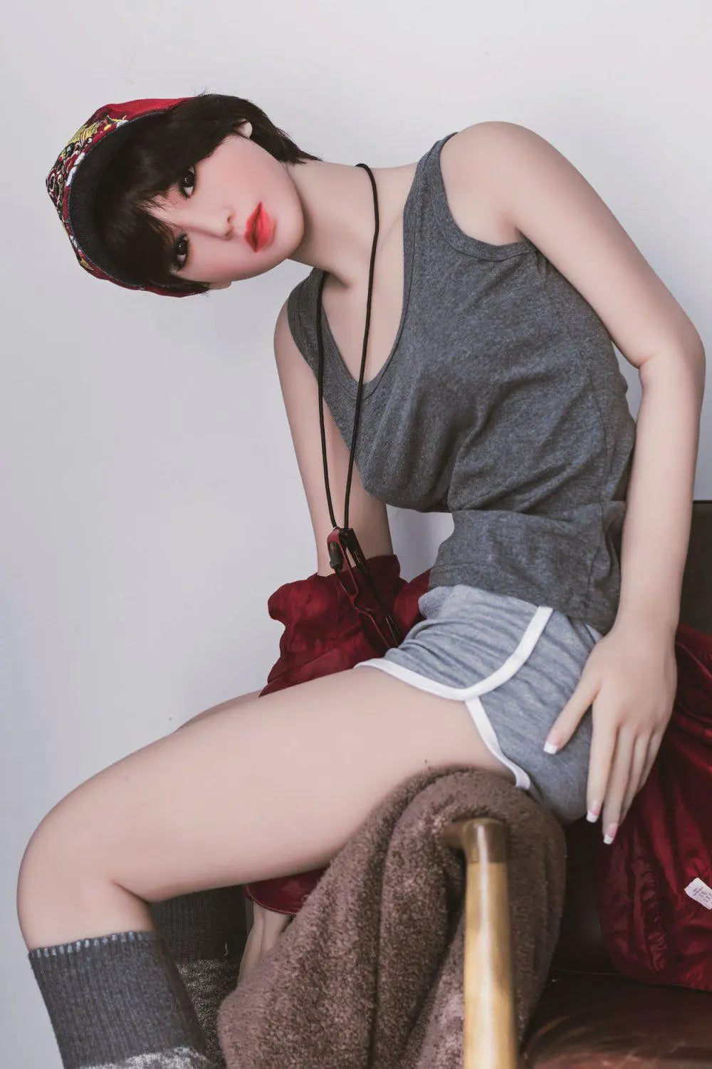 Amira short dark brown hair red lip grey tank denim shorts red jacket tomboy feminine contrast pose, sitting on the arm of the chair, baseball hat, red sunglasses,– girl-next-door companion sex doll by TopLoveDoll