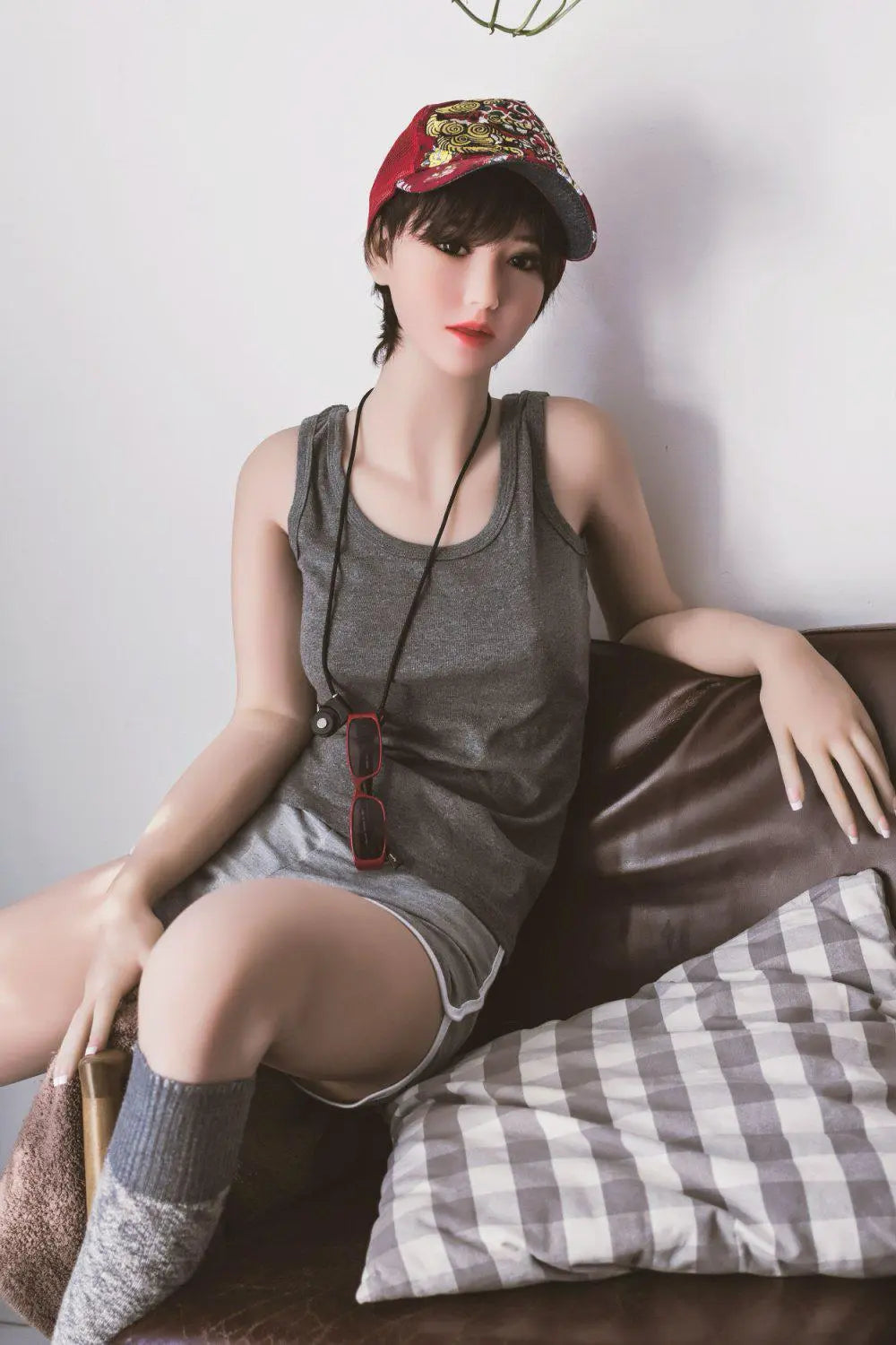 Amira short dark brown hair red lip grey tank denim shorts tomboy feminine, sitting on the arm of the chair, baseball hat, red sunglasses on the neck lace,– girl-next-door companion sex doll by TopLoveDoll