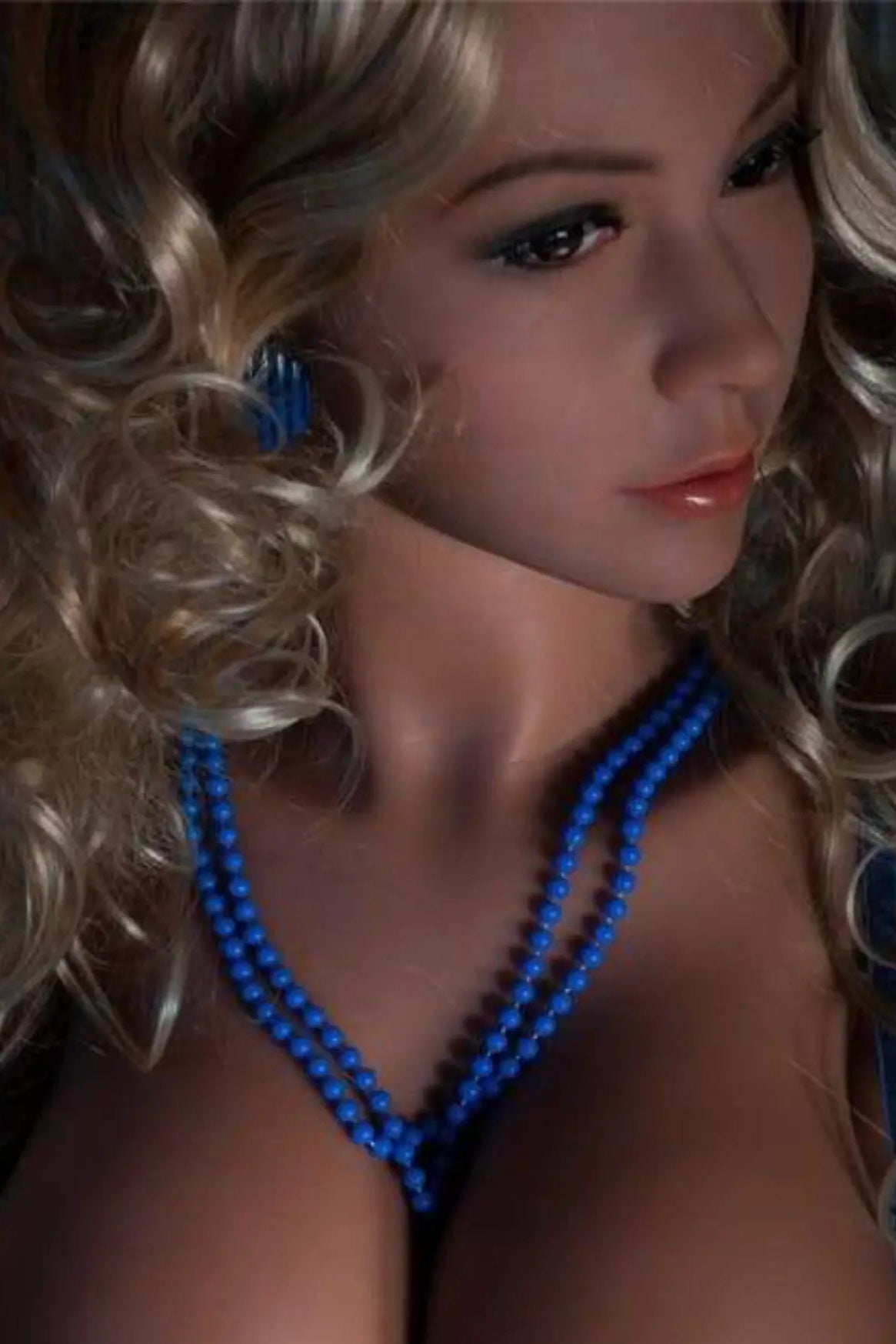 Sex doll with blonde hair and blue jewelry against a dark background